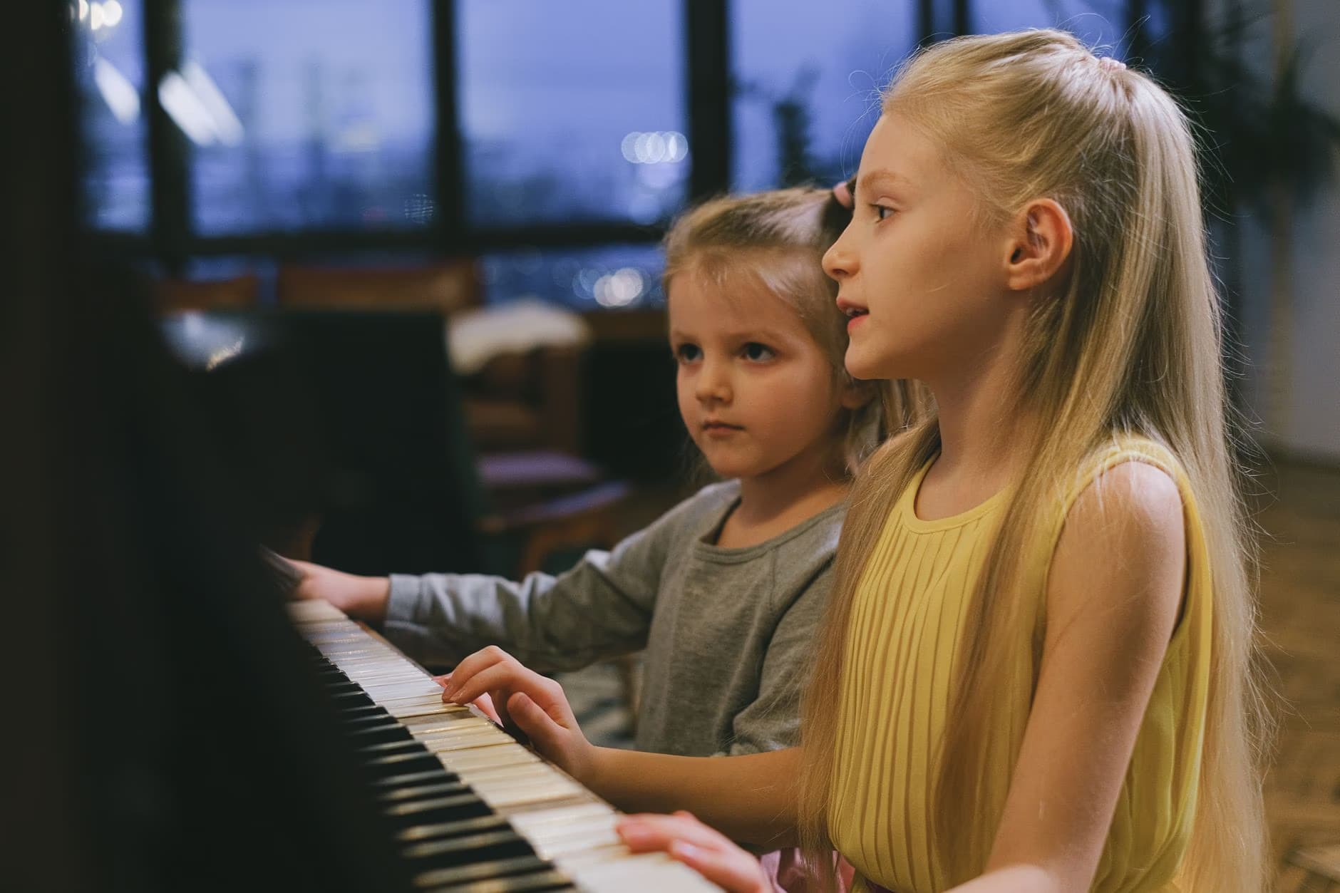 Dwie młode blondynki skupione grają razem na pianinie.