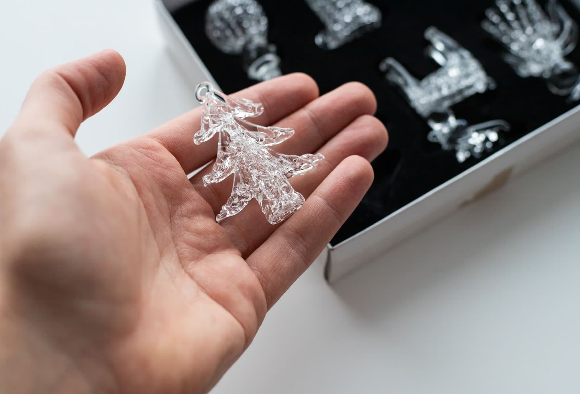 A hand holds a small, clear glass Christmas tree ornament near a box of crystal decorations.