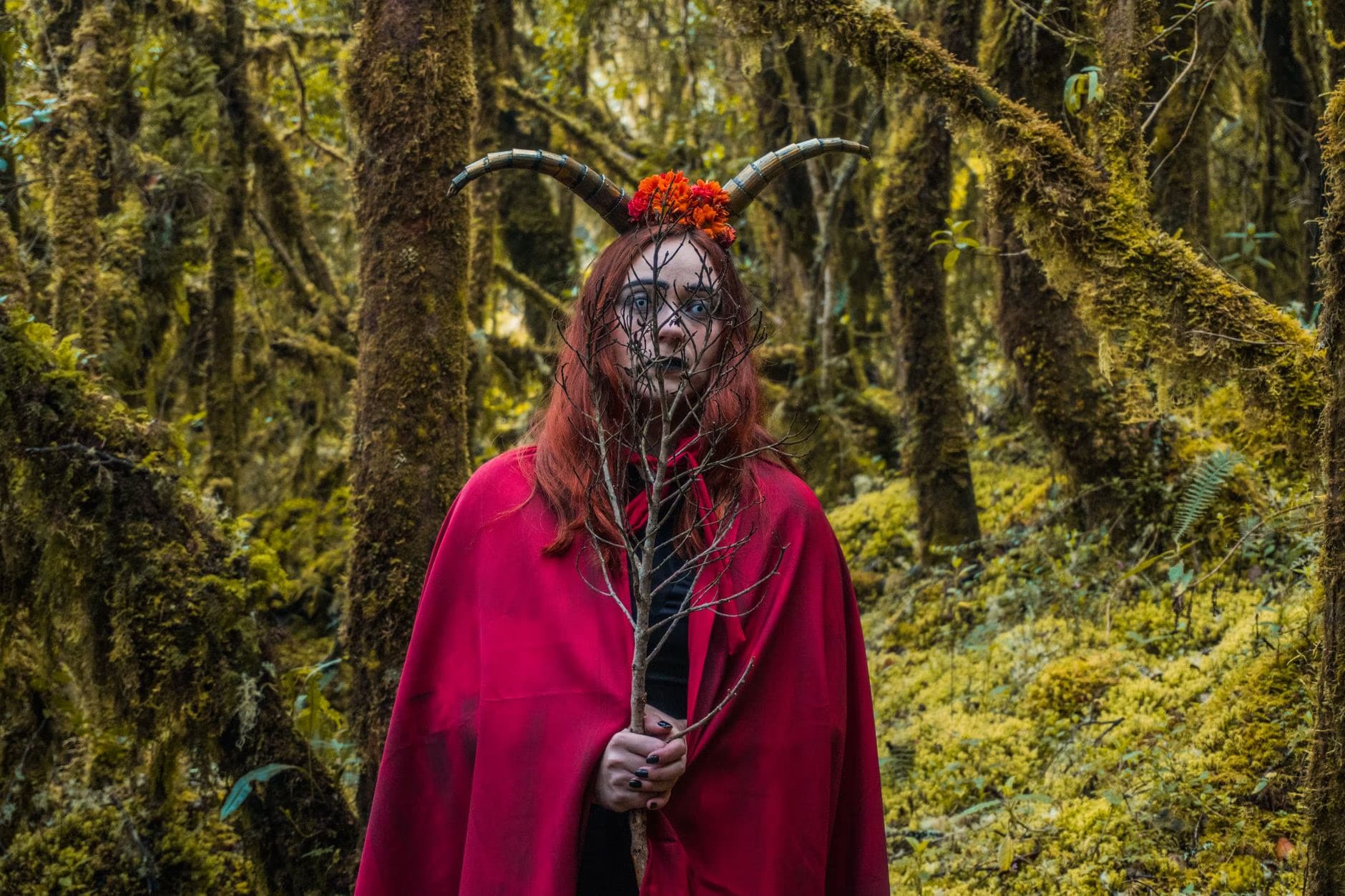 Person in a red cloak and horns posing in a mossy forest holding a branch.