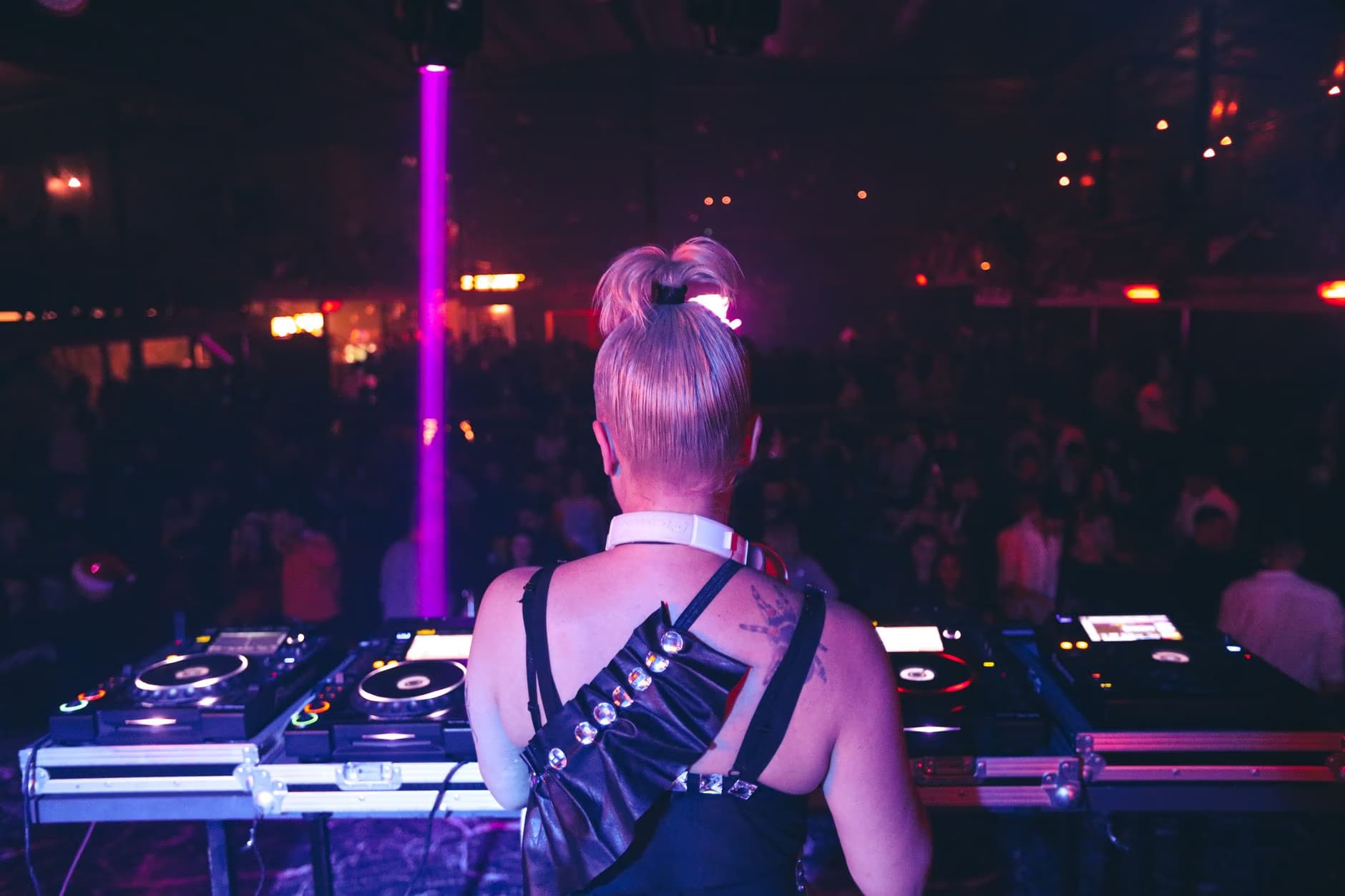 Blond DJ vystupuje za mixážním pultem v tlumeně osvětleném nočním klubu s rozmazaným davem.