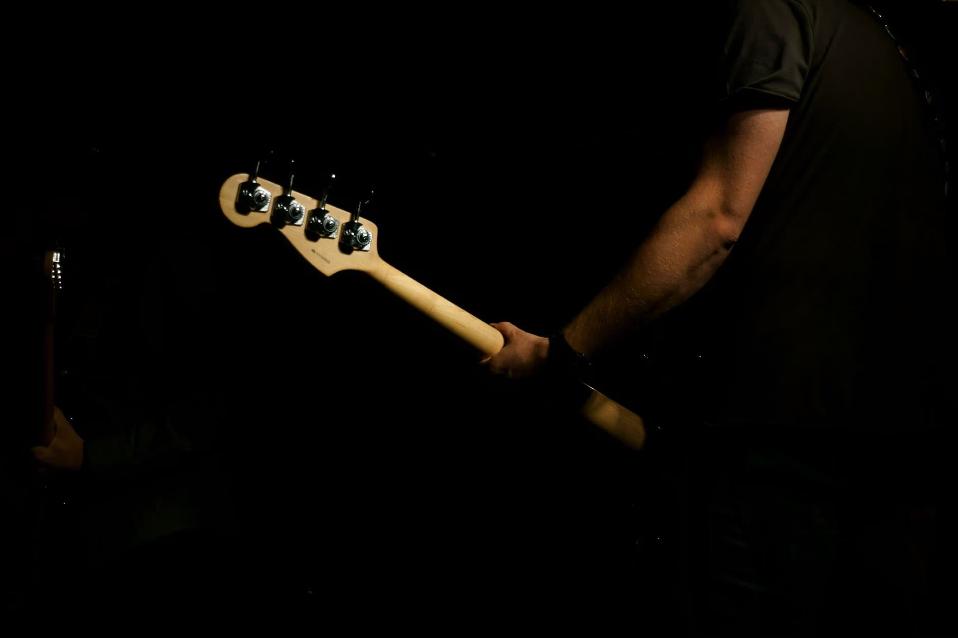Musician holding a bass guitar neck in a dimly lit, dark space.