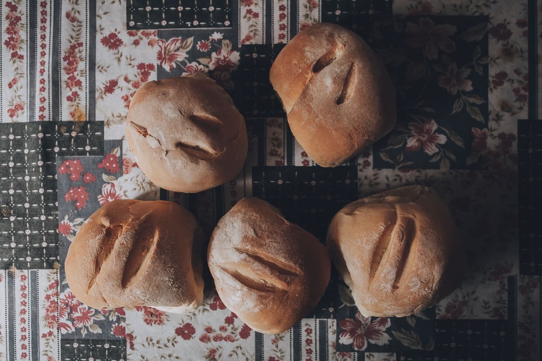Five round loaves of bread are arranged on a dark table with a floral pattern.
