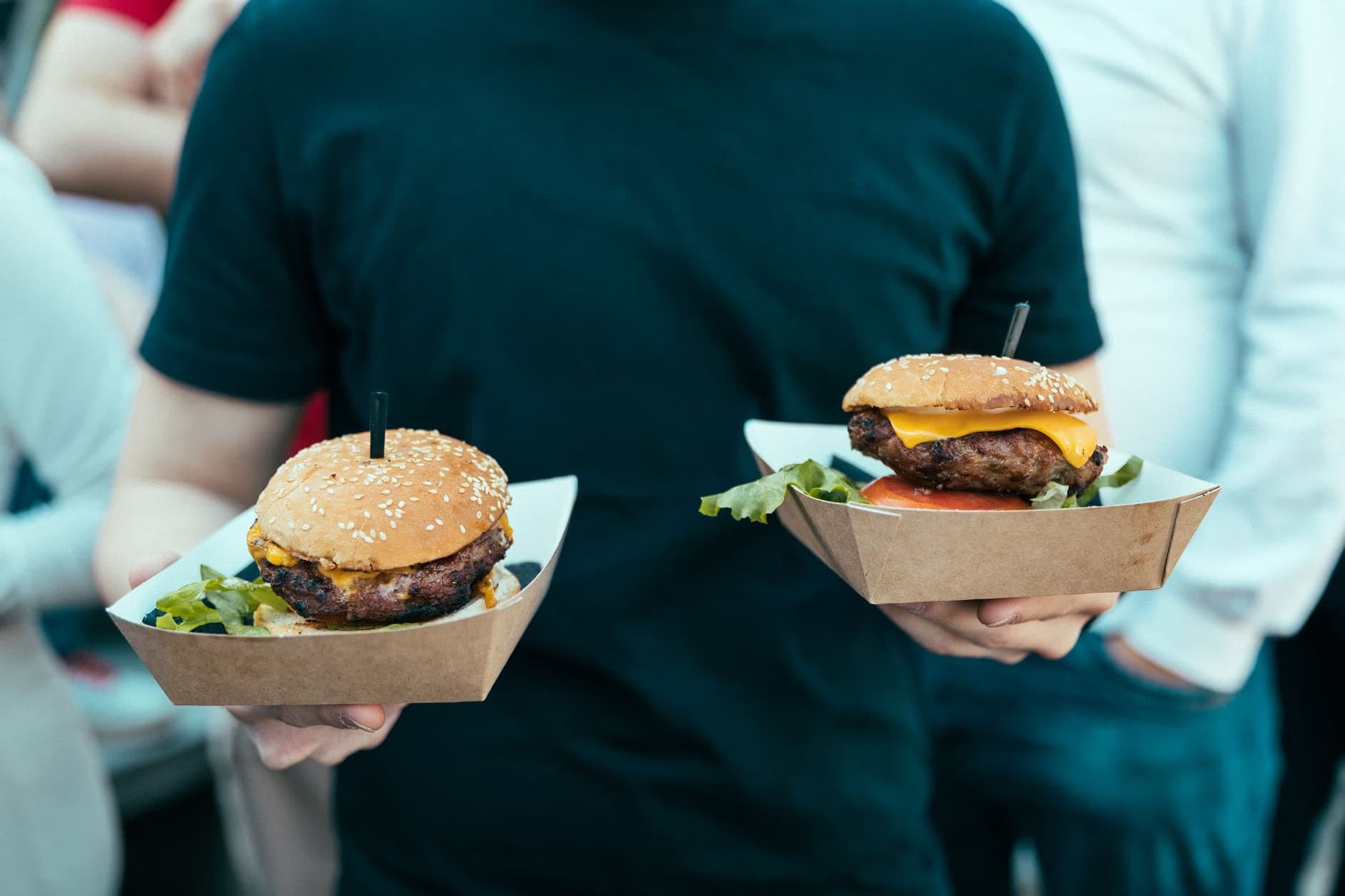 A person in a black t-shirt holds two hamburgers served in small cardboard food trays.