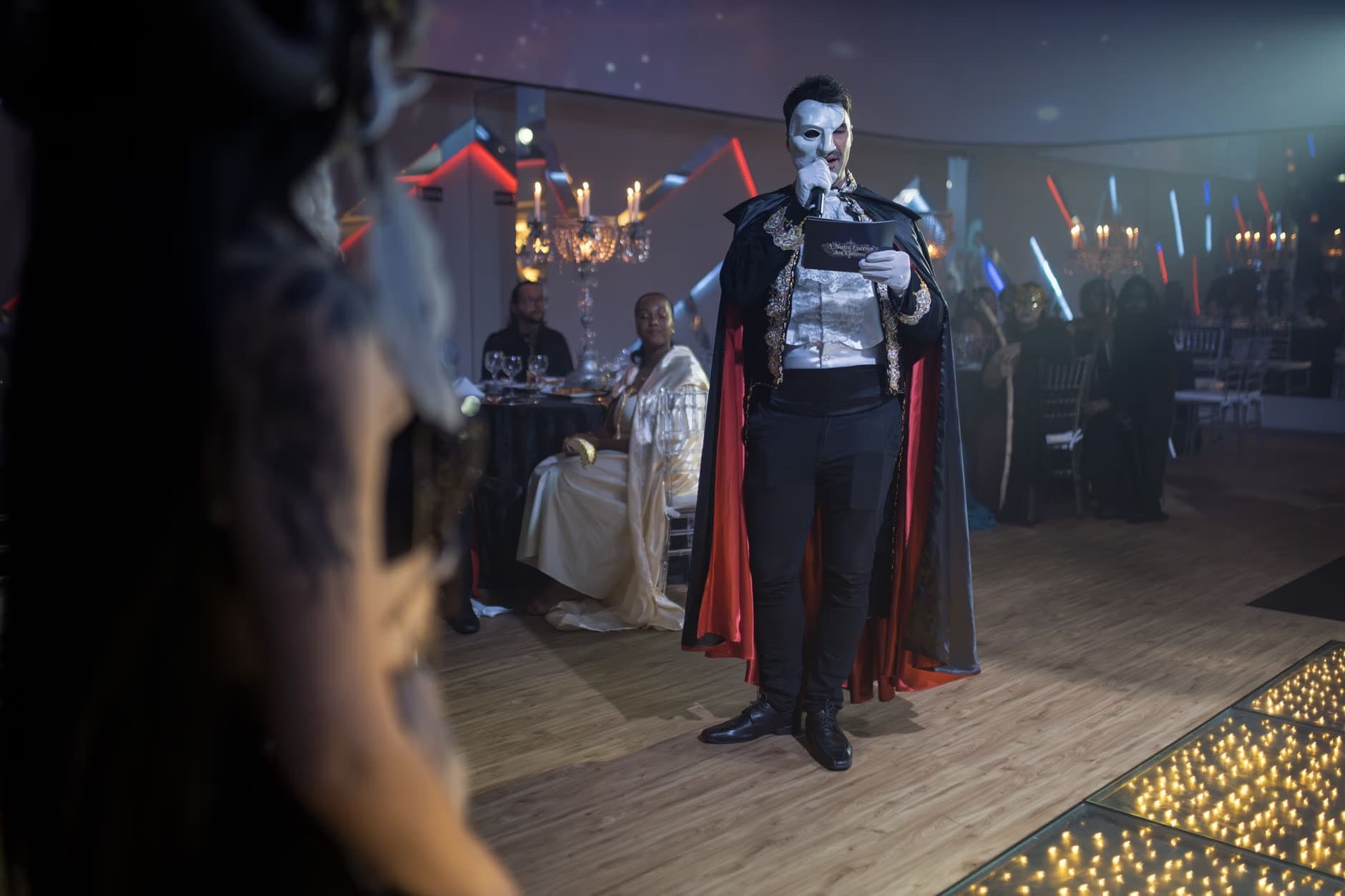 Umělec převlečený za Fantoma opery stojí na pódiu během večerní společenské akce.