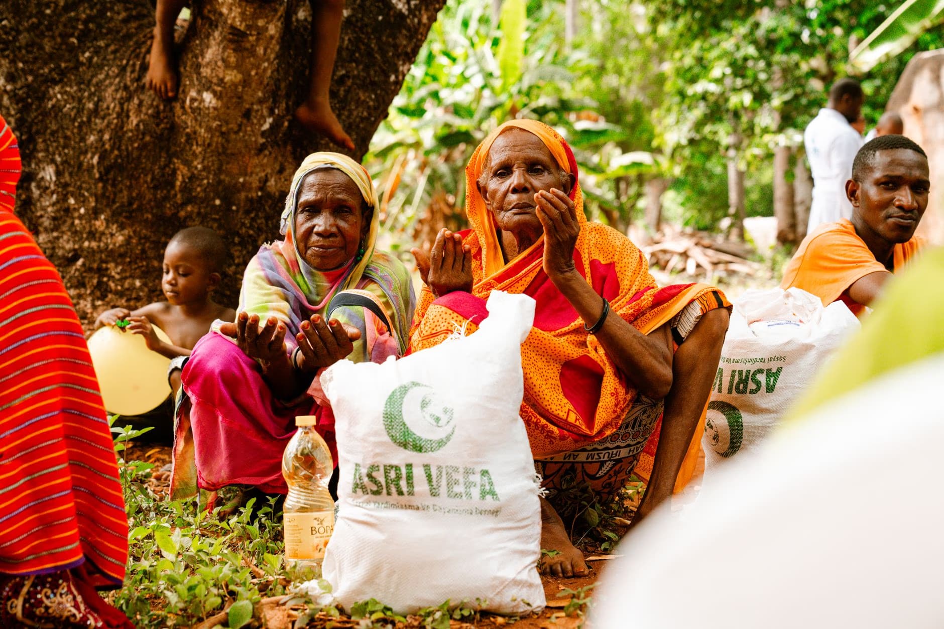 People sitting outdoors, with women wearing headscarves and holding bags marked Asri Vefa.