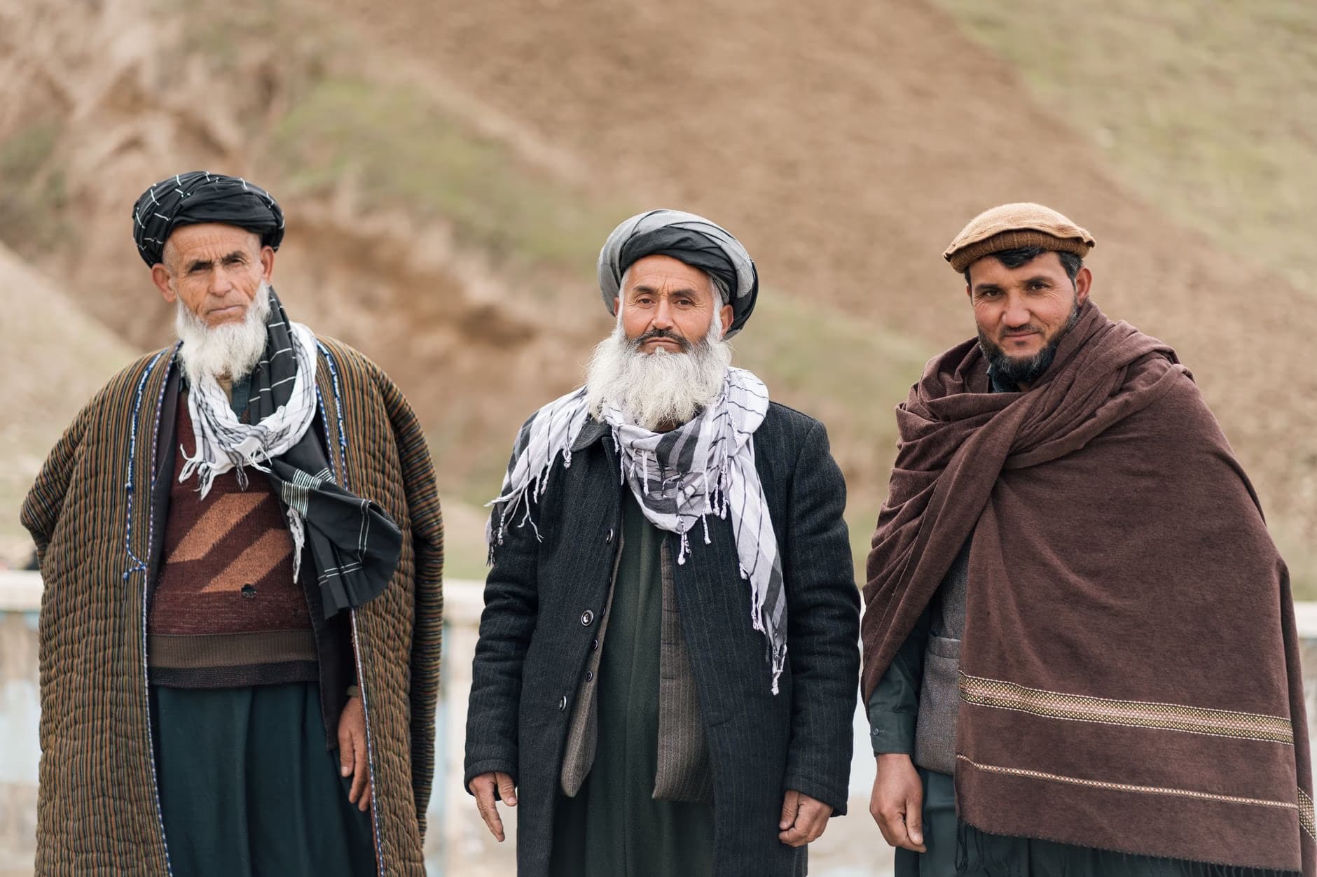 Three men with traditional Afghan attire and beards stand together outdoors in a landscape.