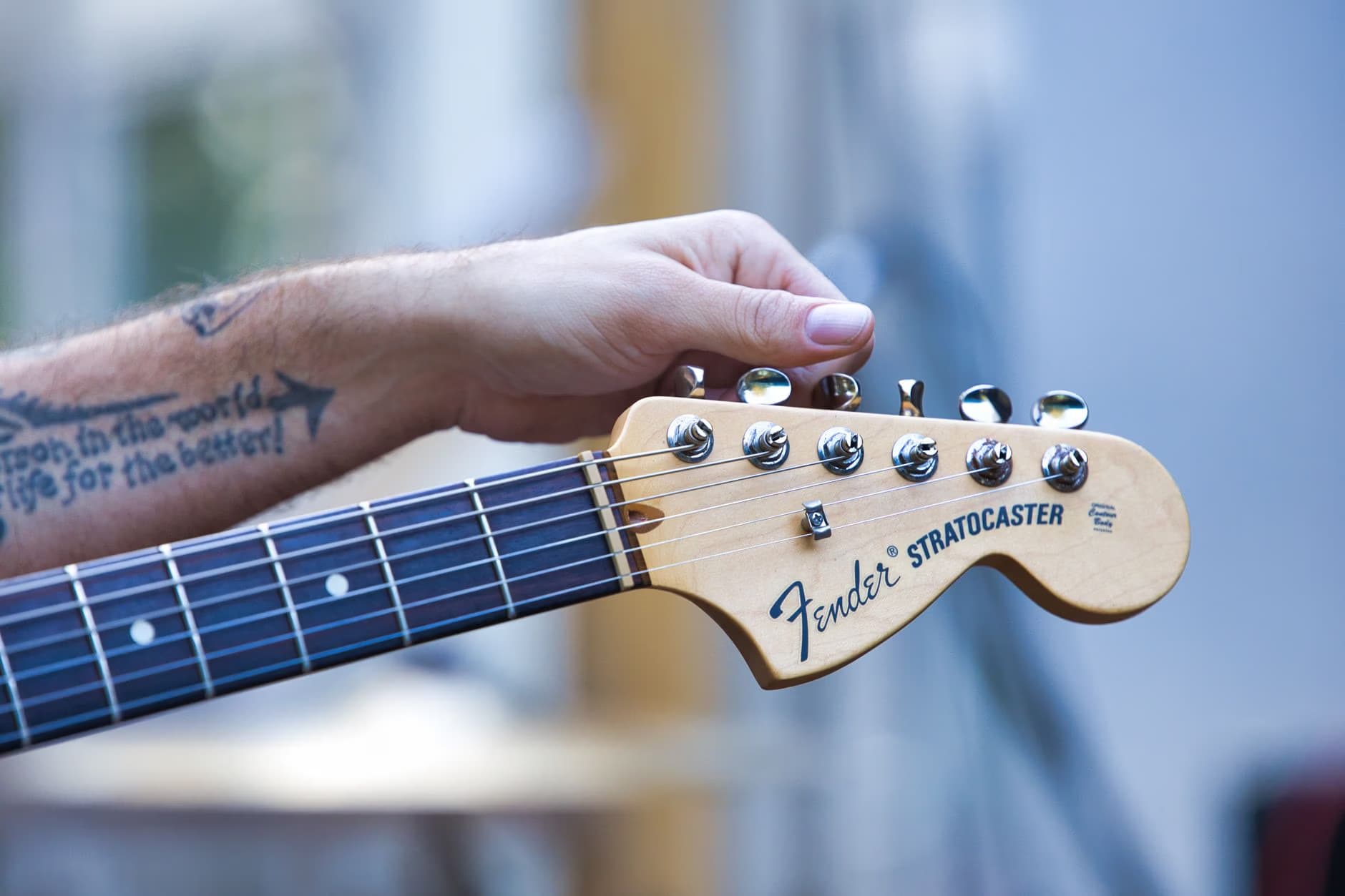 A close-up of a musician's hand tuning a Fender Stratocaster guitar headstock.
