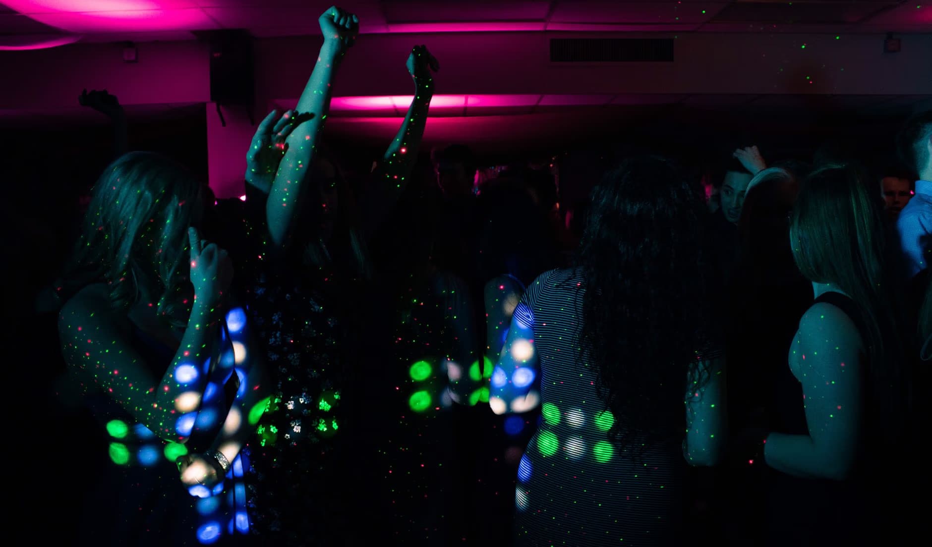 People dance in a dimly lit nightclub illuminated by vibrant pink and blue laser lights.