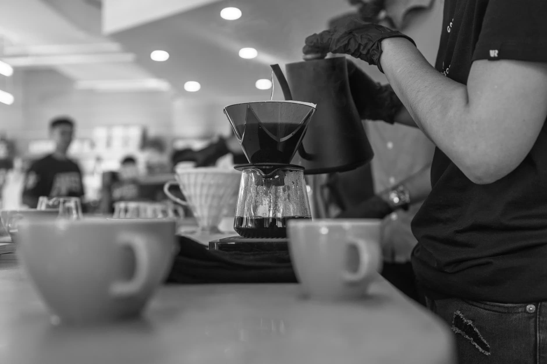 A barista in black gloves pours hot water from a kettle into a coffee dripper at a shop counter.