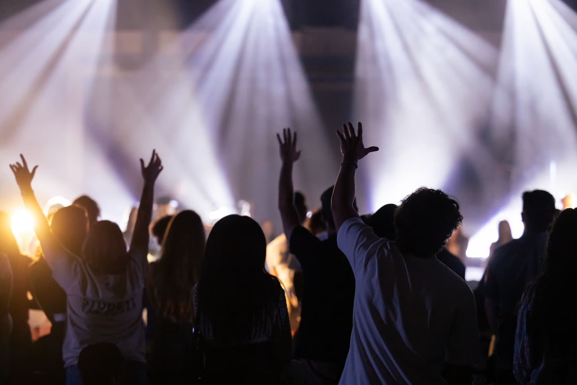 Silhouettes of people with raised hands at a concert under bright stage lights.