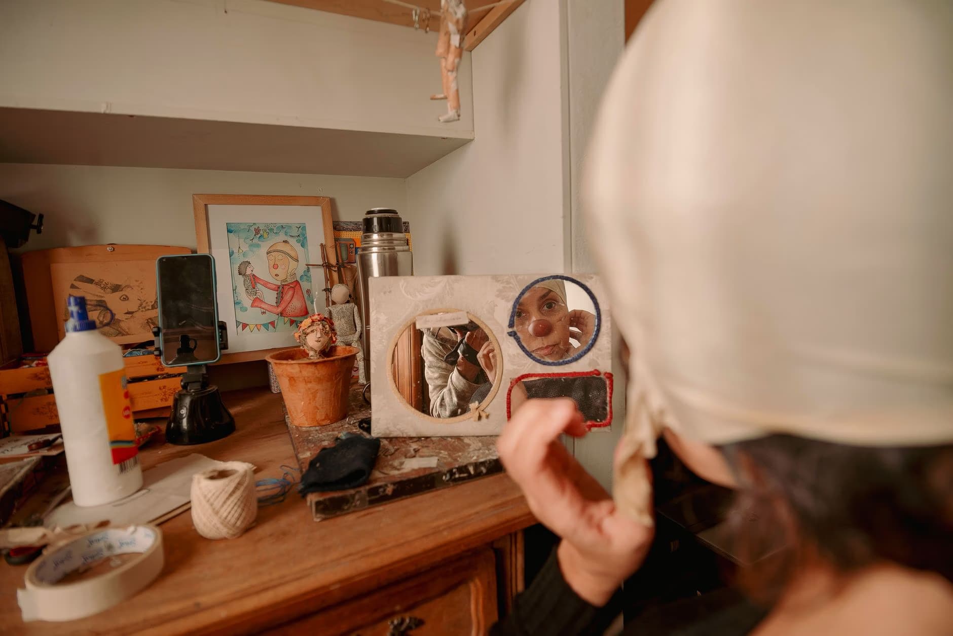A person with a white head covering looks into a small mirror on a cluttered workbench.