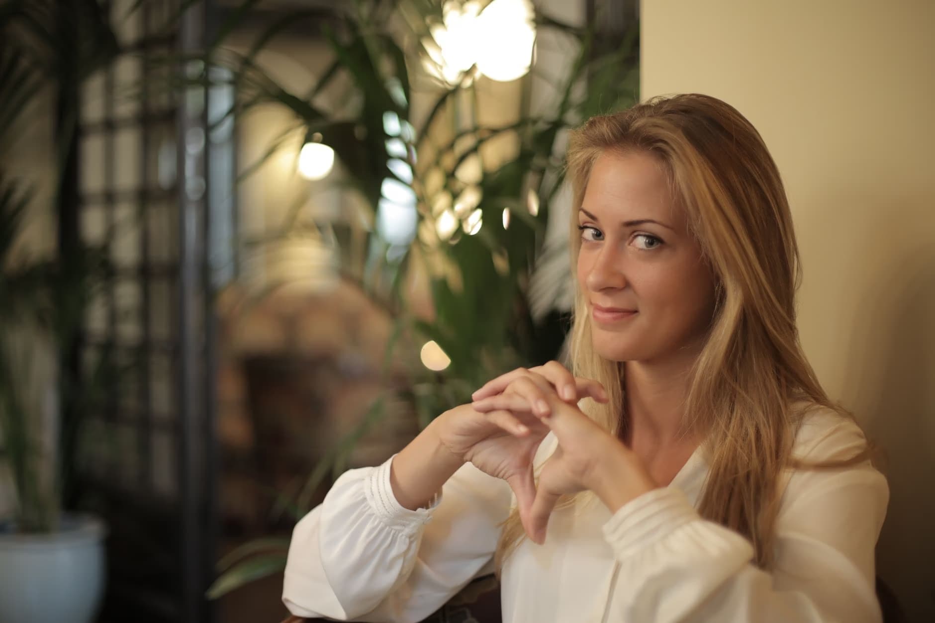Blonde woman with long hair smiles softly while sitting near green potted plants indoors.