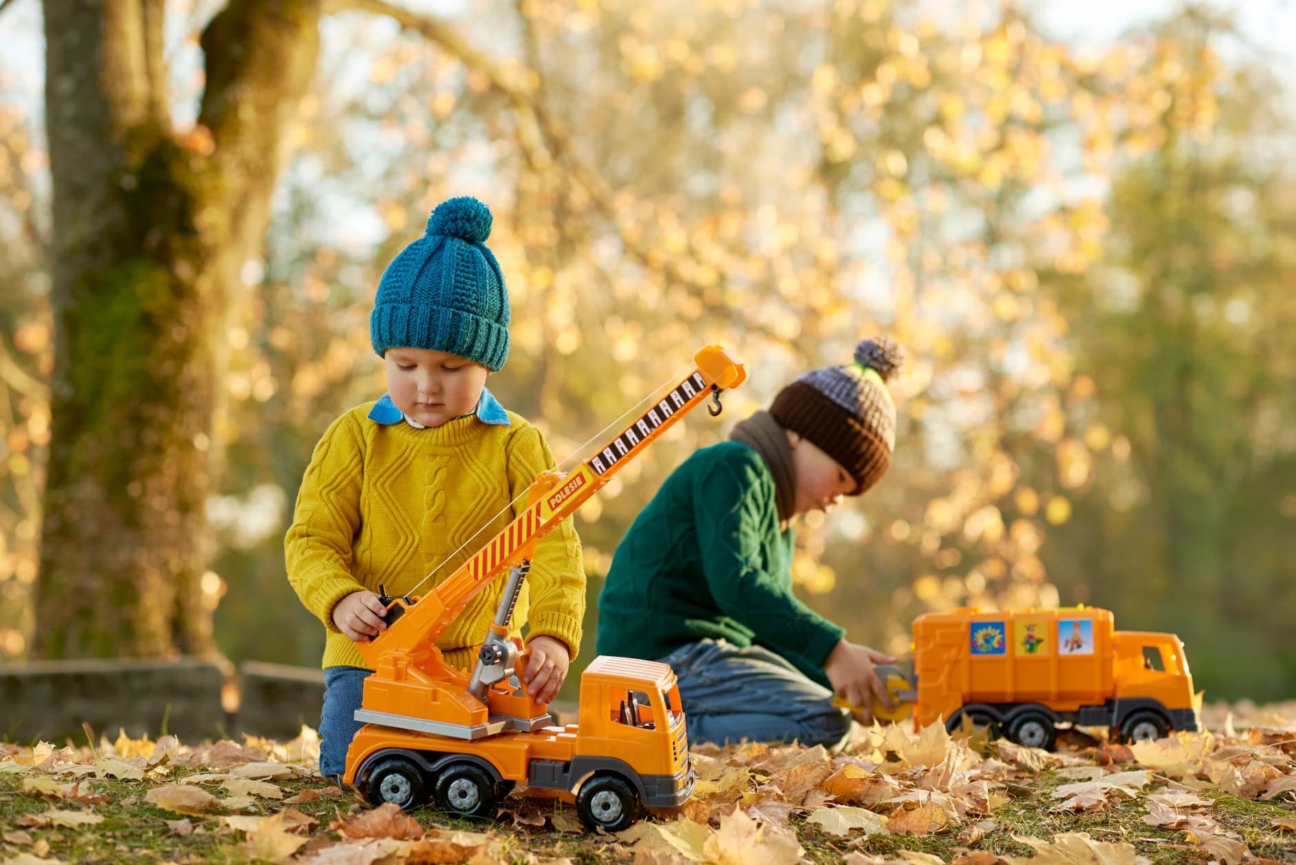 Two young children in warm hats play with toy construction trucks in a park filled with autumn leaves.