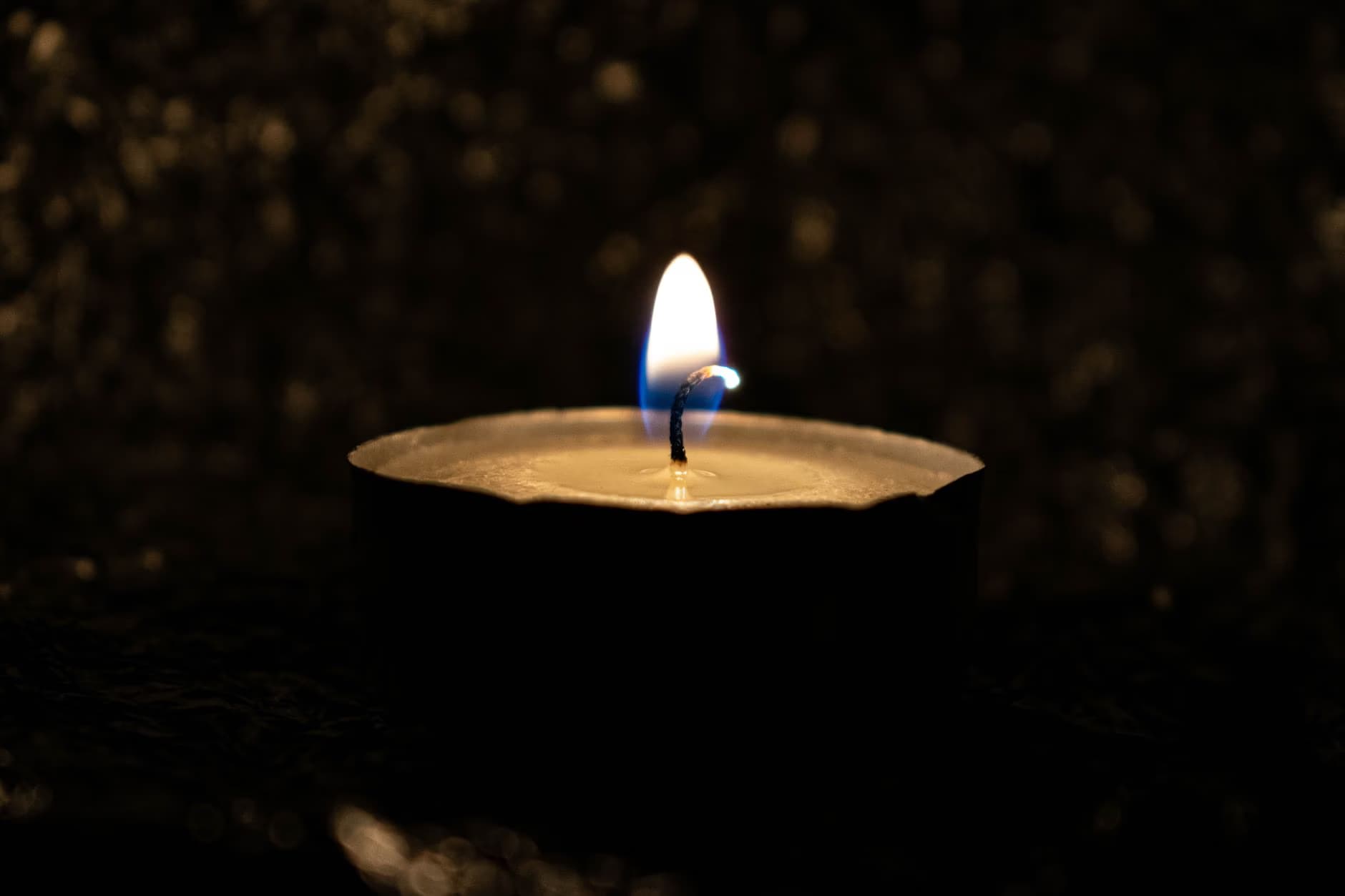 Ein einzelnes brennendes Teelicht leuchtet hell vor einem dunklen, unscharfen Hintergrund.