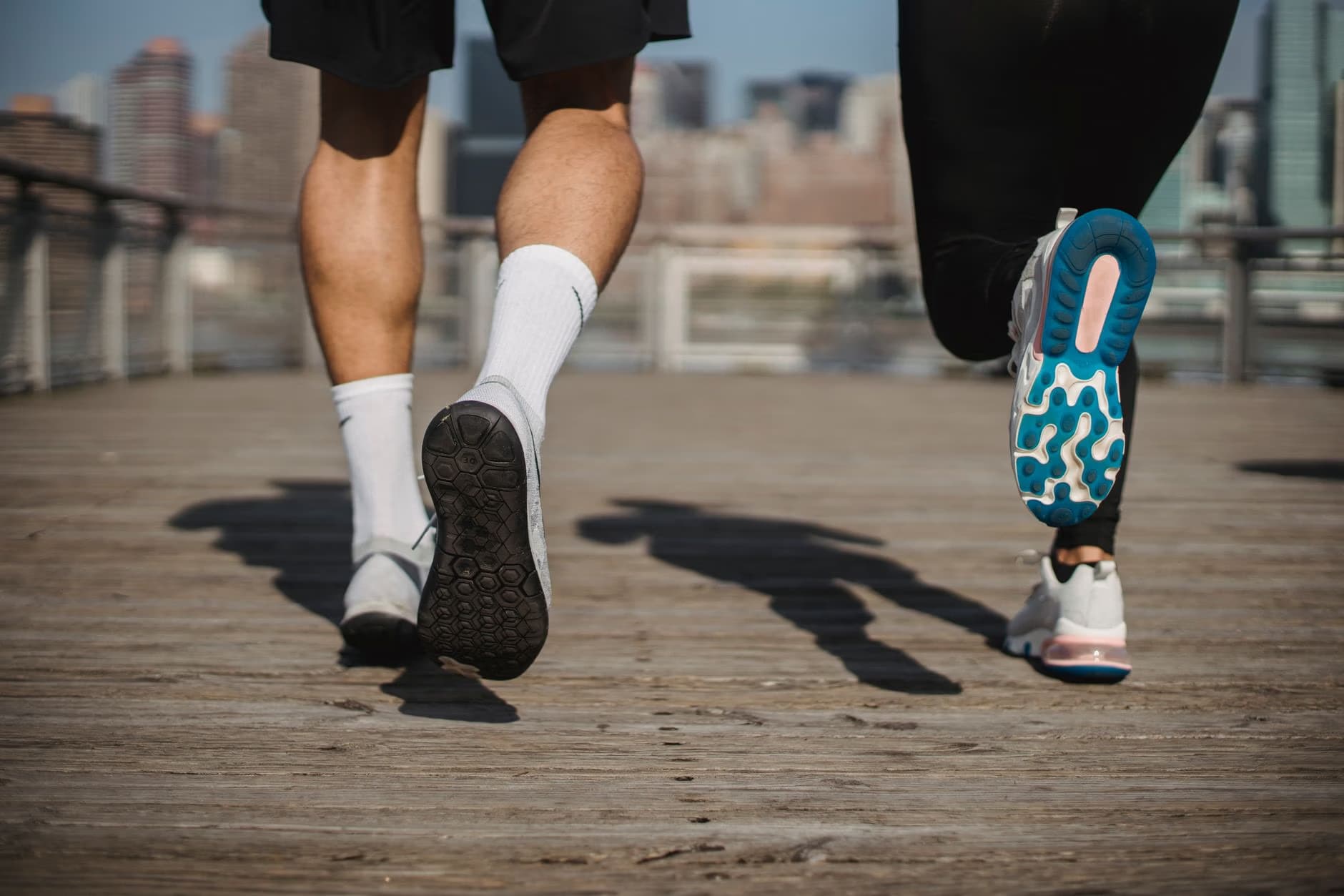Two runners in sportswear jog on a wooden waterfront promenade with a blurry city skyline.