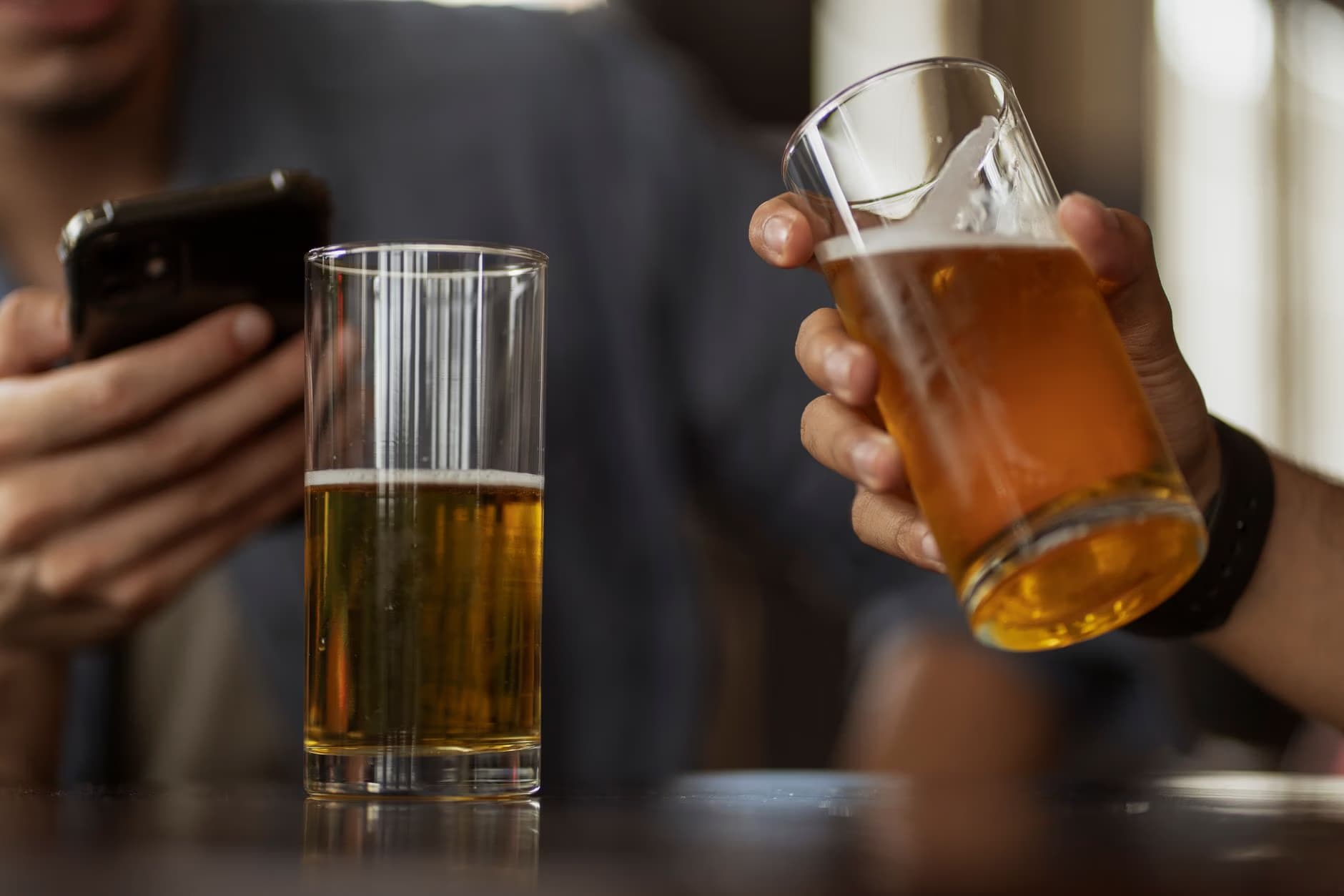 Two people hold glasses of beer while one person looks at a mobile phone.