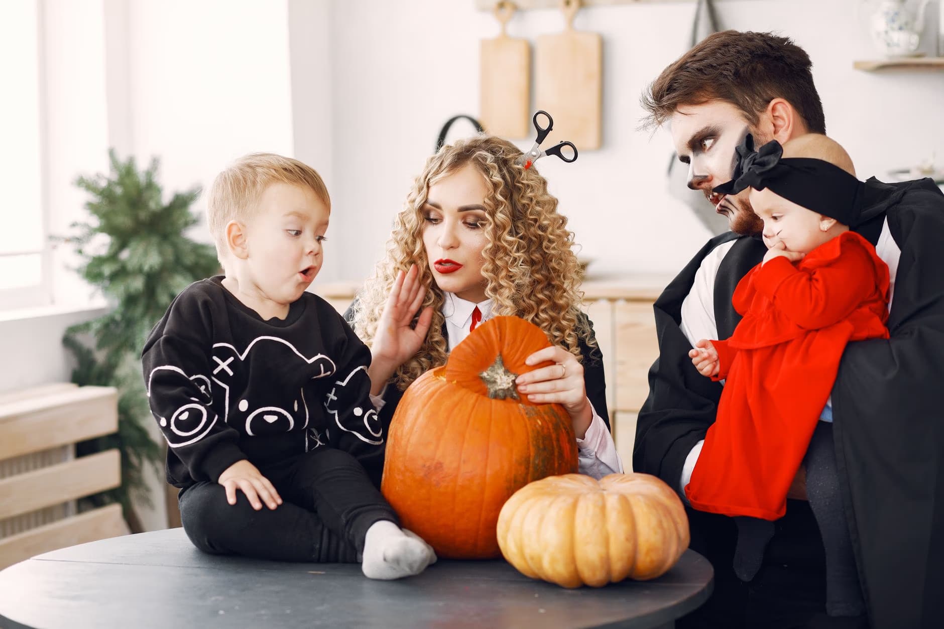 Rodina v halloweenských kostýmech sedí u stolu s dýněmi ve světlé místnosti.