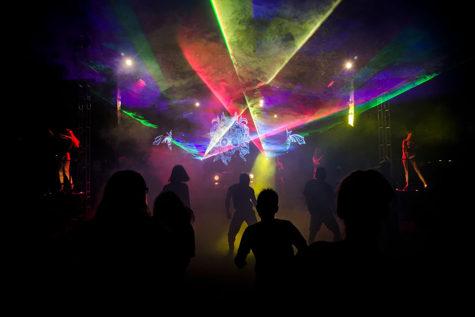 Silhouettes of dancers move in a dark club under vibrant, smoky laser beams.