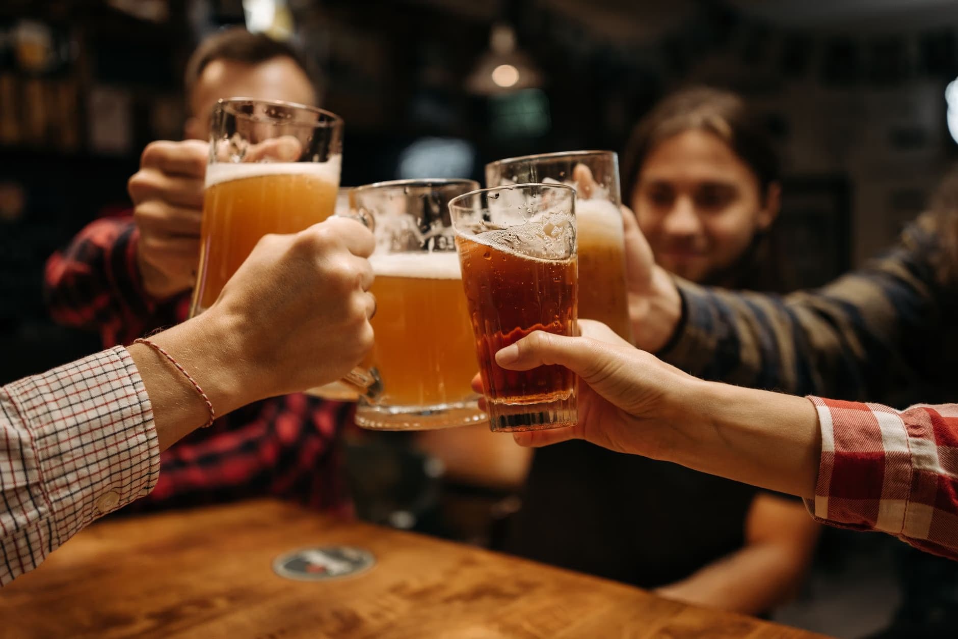 Friends clink beer mugs together over a wooden pub table.