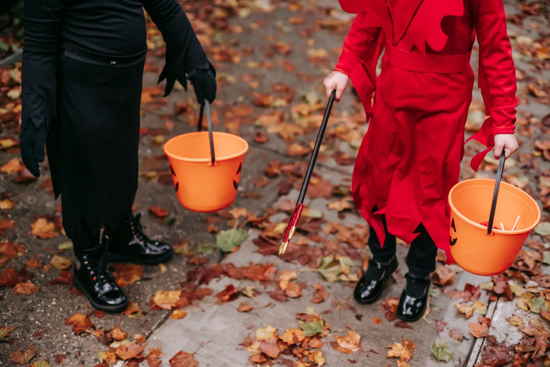 Two children in Halloween costumes hold orange candy buckets while walking on fallen autumn leaves.
