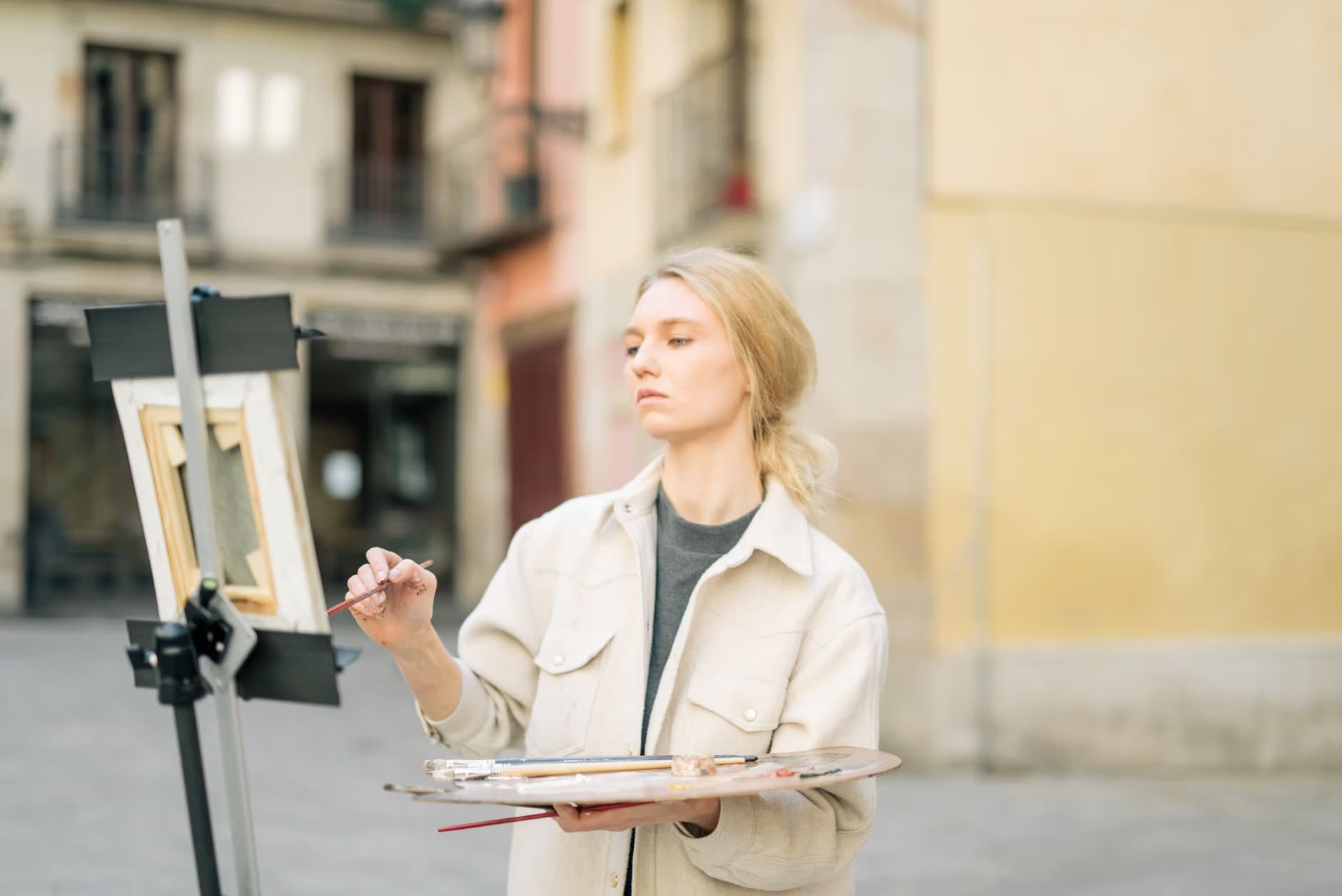 A young blonde woman paints on a canvas easel while holding a wooden palette in a city square.