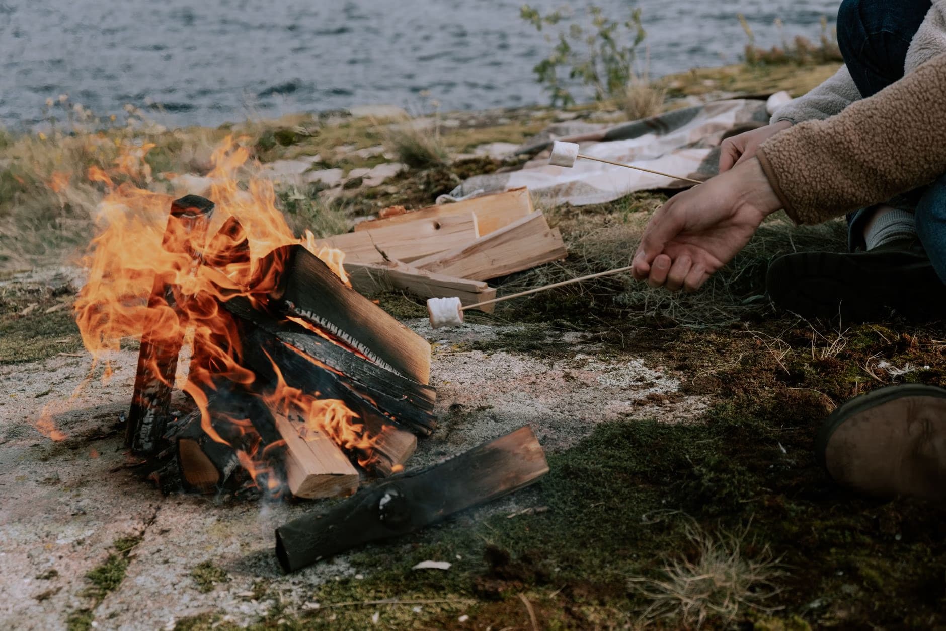Osoba piecze piankę marshmallow nad małym ogniskiem na skalistym brzegu jeziora.