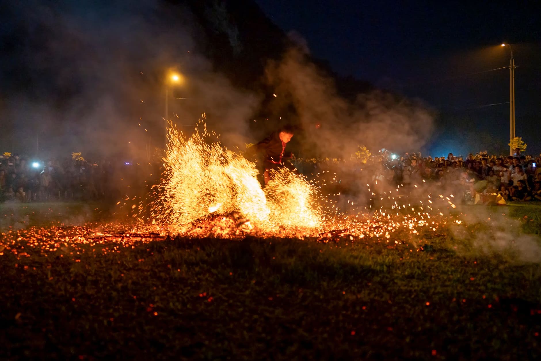 A large, glowing bonfire burns brightly in a dark field at night, surrounded by scattered sparks.