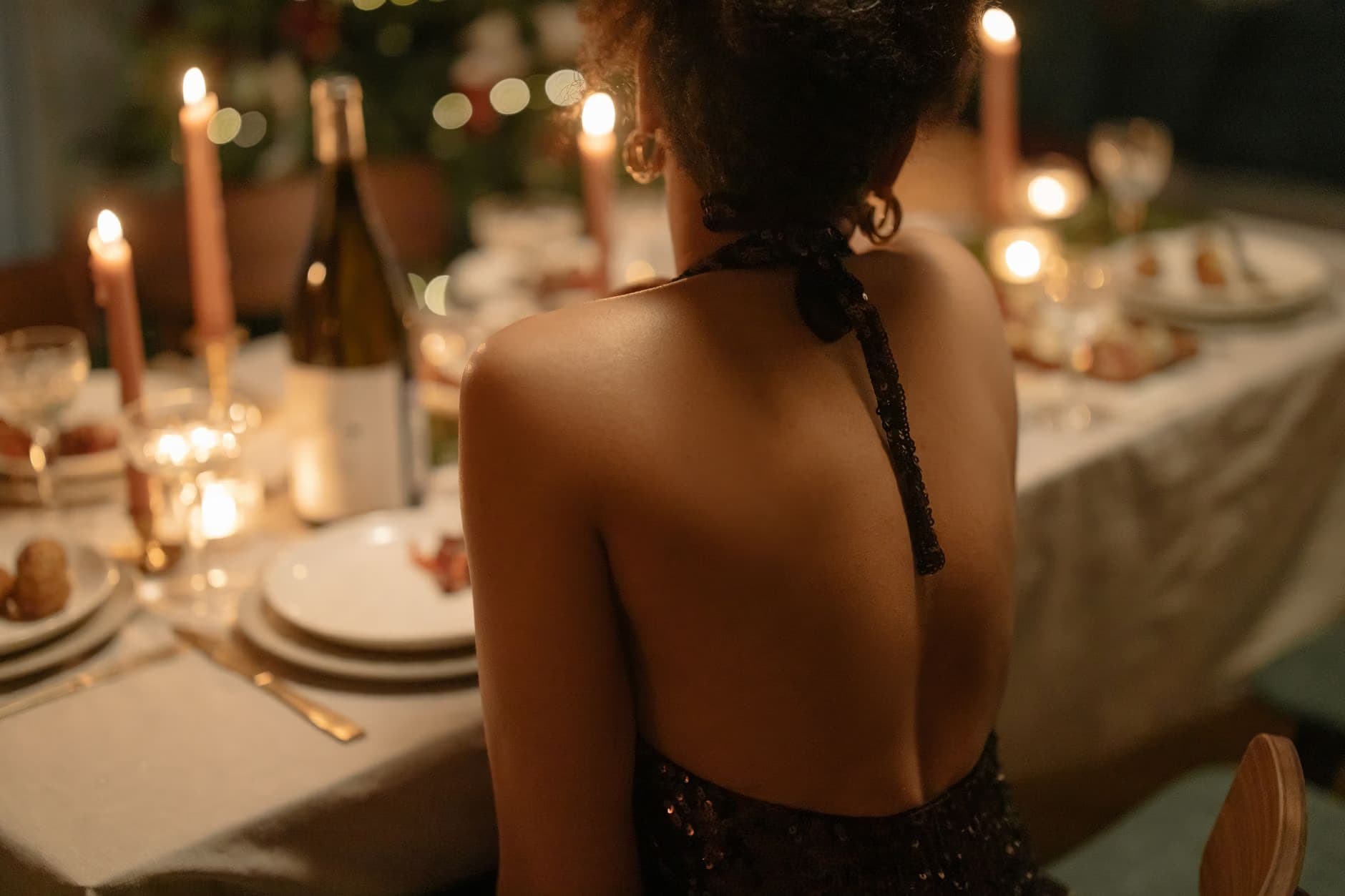 A woman in a backless evening dress sits at a candlelit dinner table with wine.