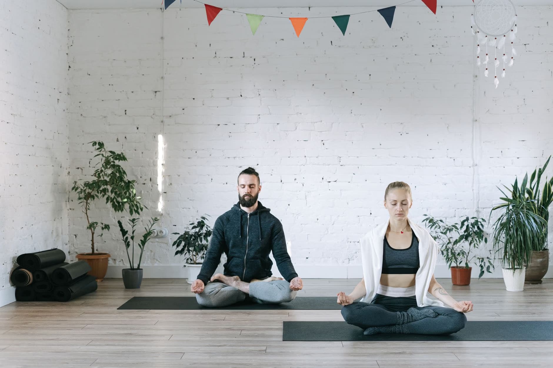 Ein Mann und eine Frau meditieren auf Yogamatten in einem hellen Studio mit Pflanzen und Wimpeln.