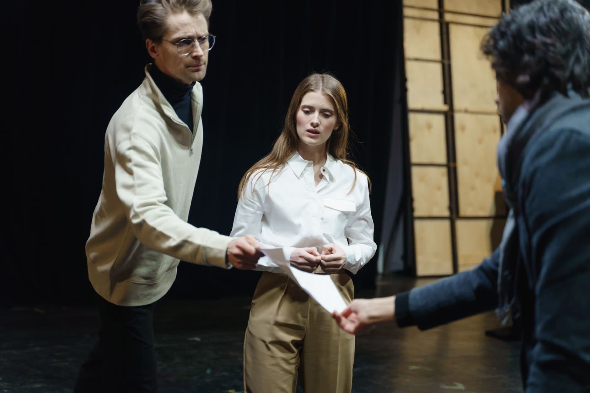 Three actors in casual clothing rehearse a scene on a dimly lit theater stage.