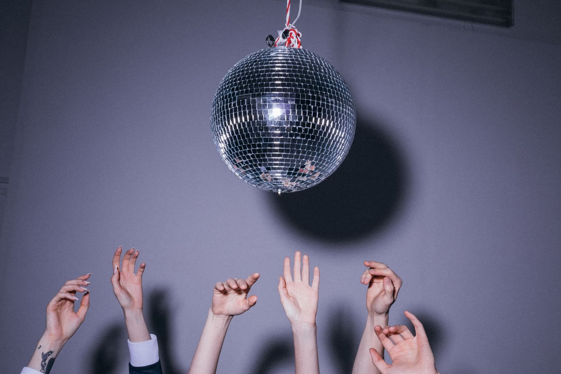 Eine glänzende Discokugel hängt in einem dunklen Raum über mehreren Menschen mit erhobenen Händen.