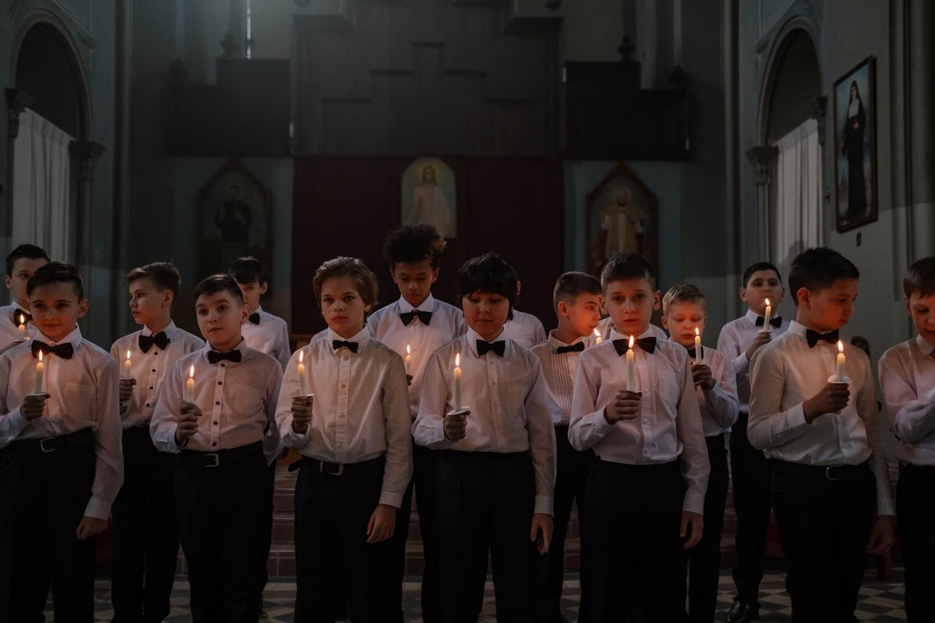 Grupa chłopców w białych koszulach i muchach trzyma zapalone świece wewnątrz kościoła.