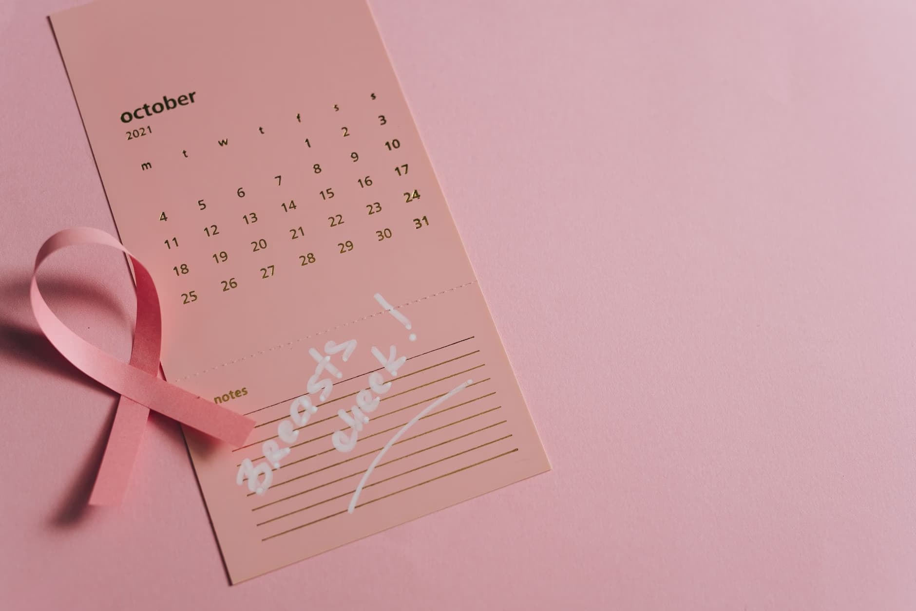 A pink ribbon lies next to a calendar for October 2021 with the words Breast check written on it.