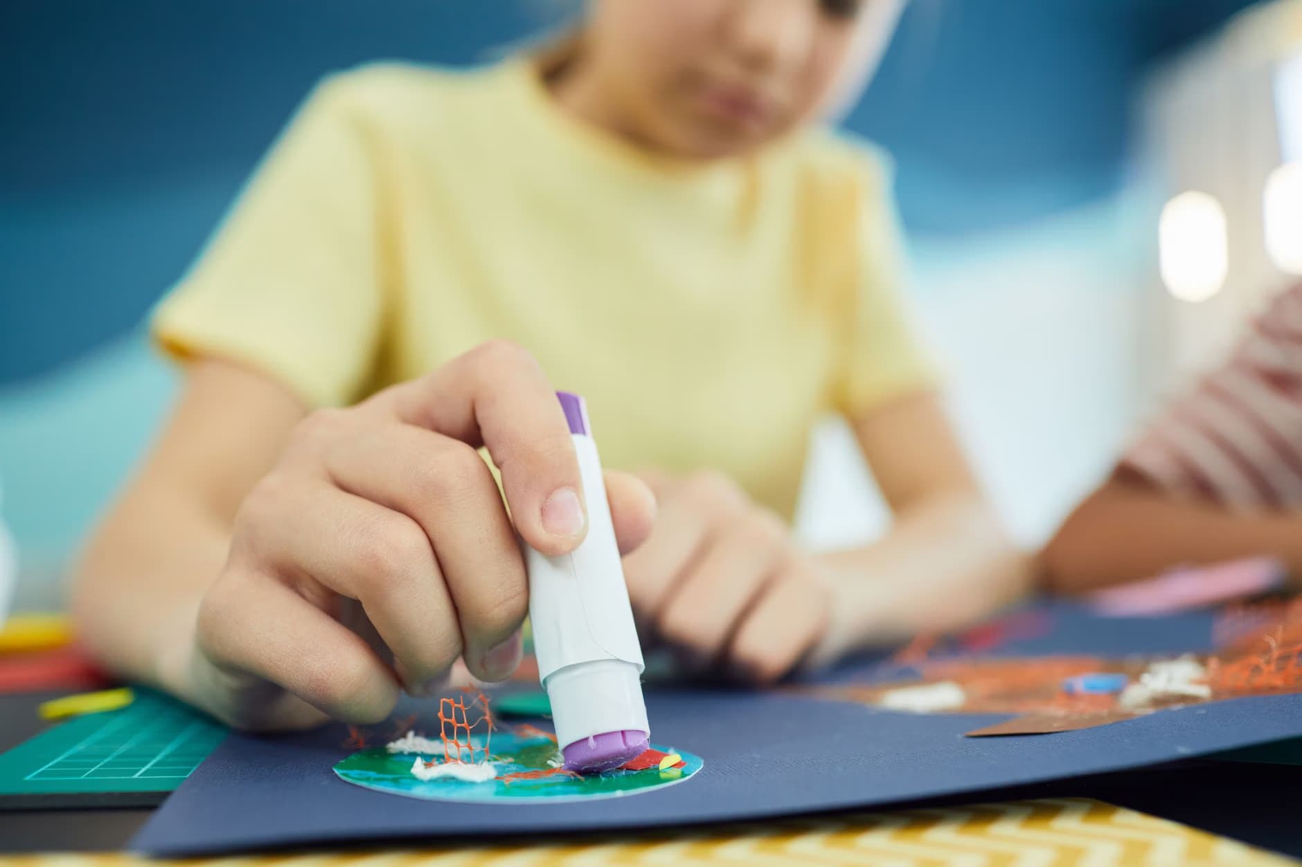 A child in a yellow shirt uses a glue stick to craft on blue paper.