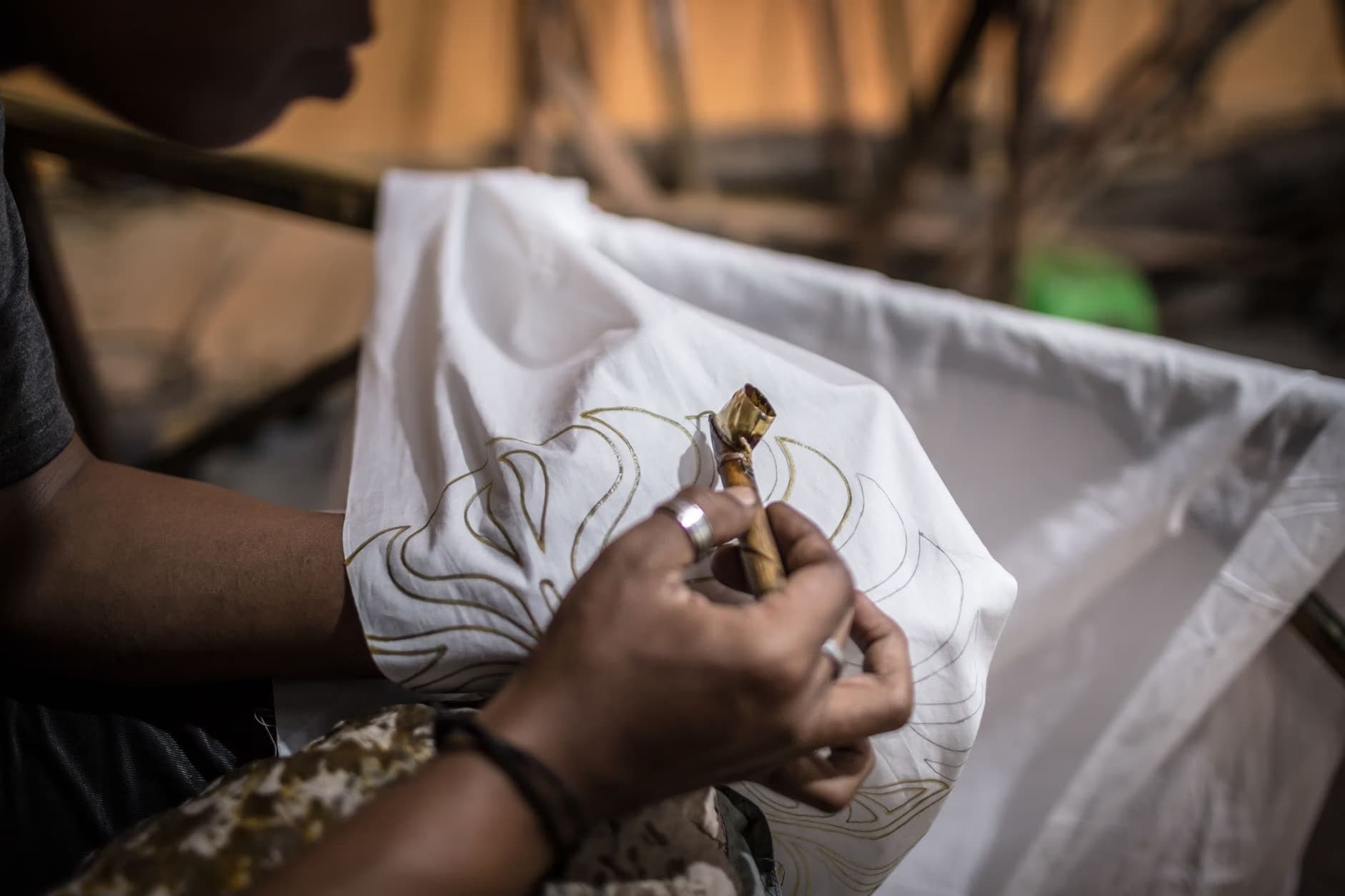 A close-up shot of a person using a traditional canting tool to apply wax patterns onto white fabric.