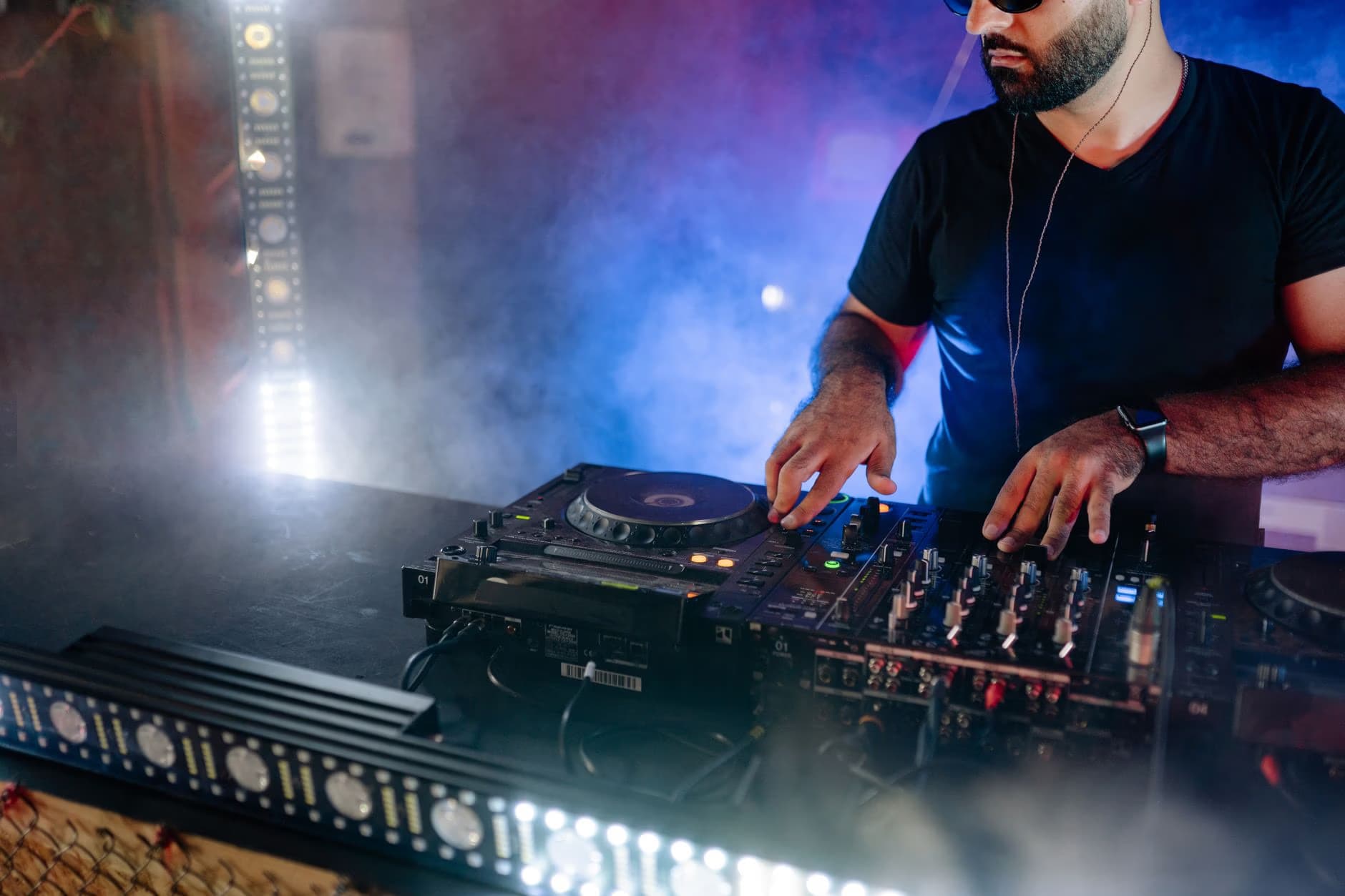 Brodaty DJ w czarnym t-shircie gra muzykę na profesjonalnym sprzęcie w zadymionym klubie nocnym.