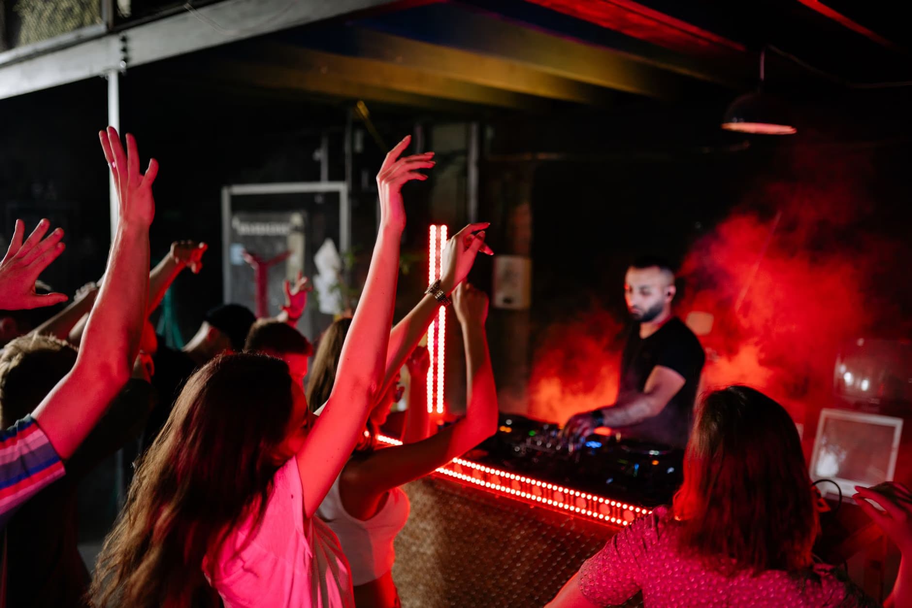Lidé tančí v temném klubu s červeným osvětlením, zatímco DJ pouští hudbu za mixážním pultem.