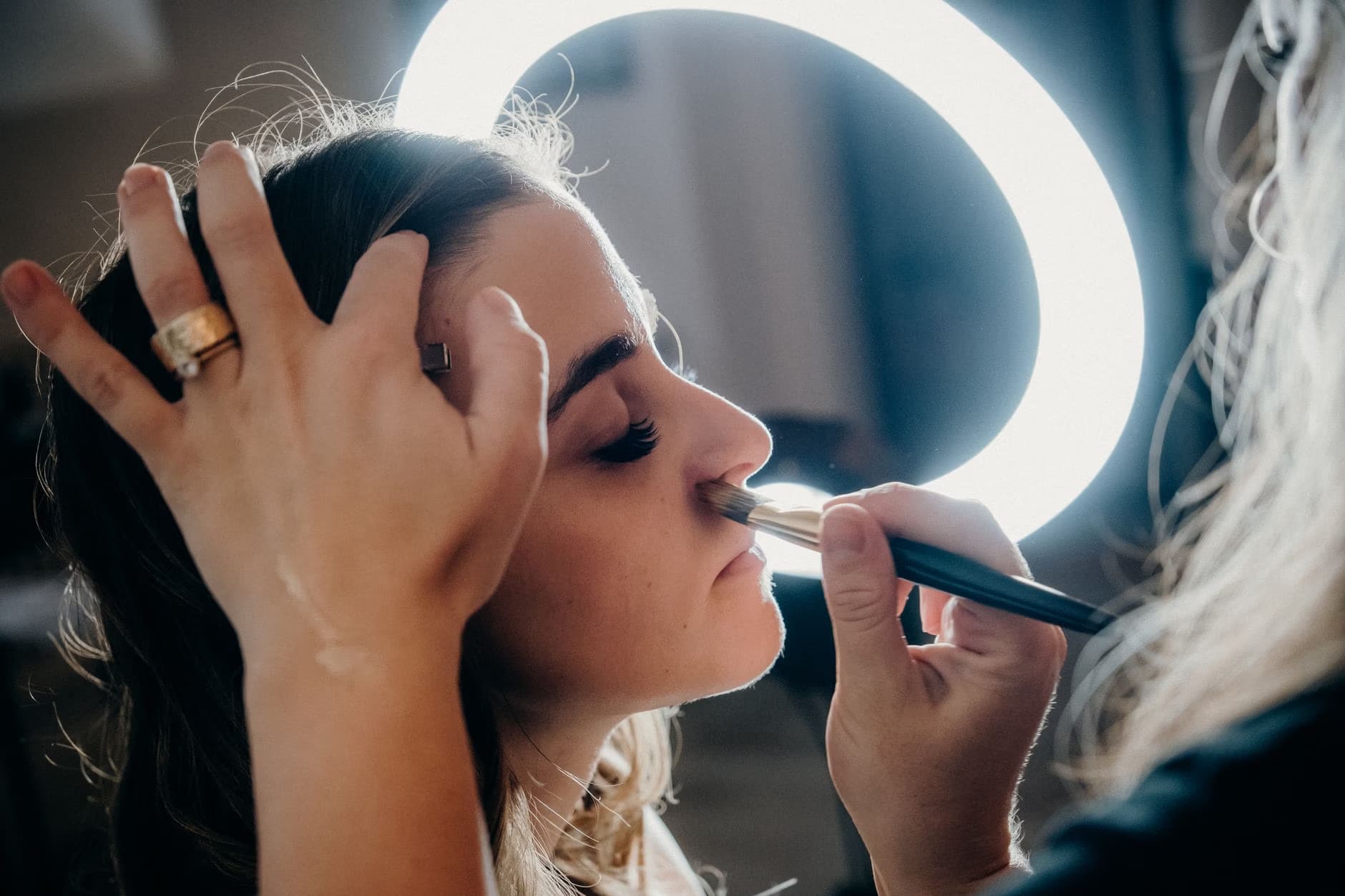 Makeup artist applies makeup to a client's face under bright ring light illumination.