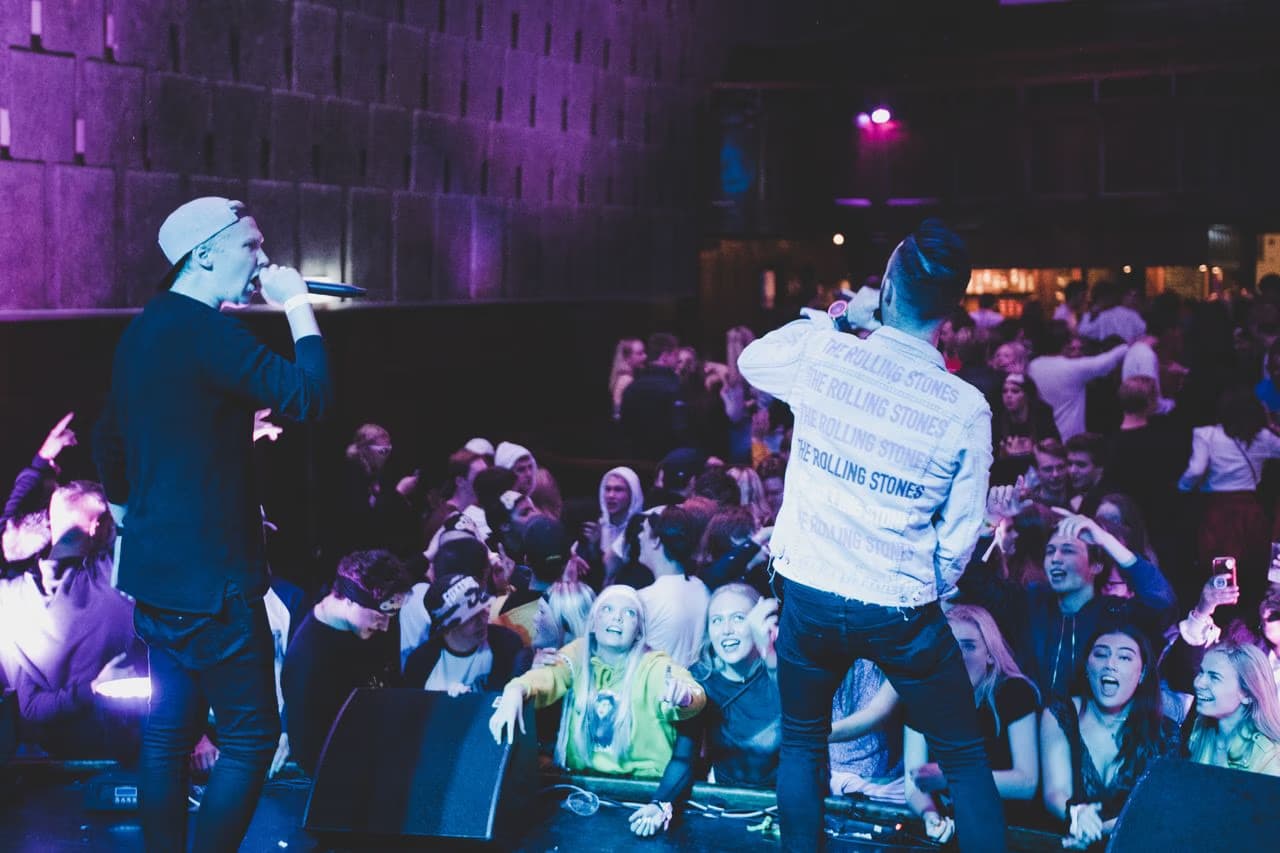 Two performers hold microphones on stage in front of a cheering crowd at a club event.