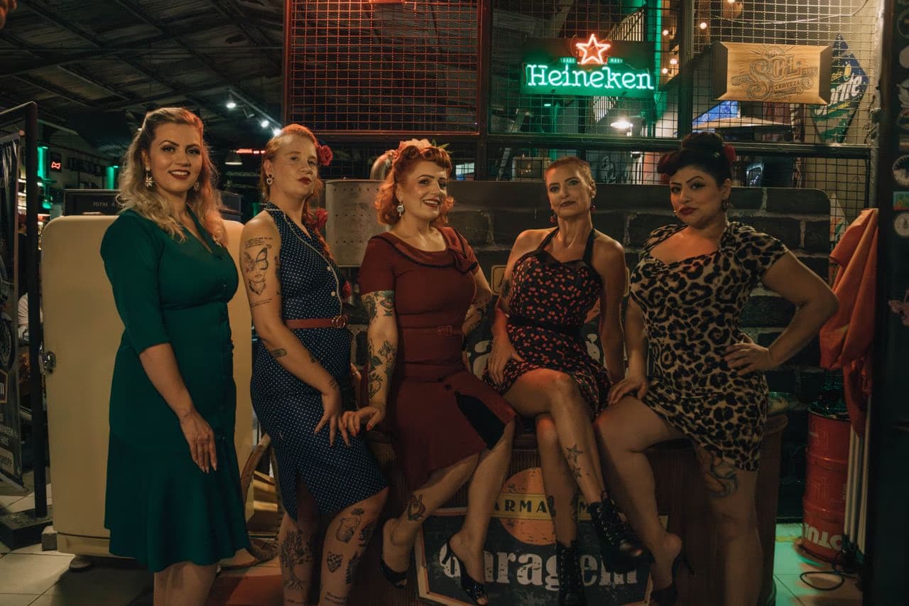 Five women pose in vintage style clothing inside a bar with a neon Heineken sign.