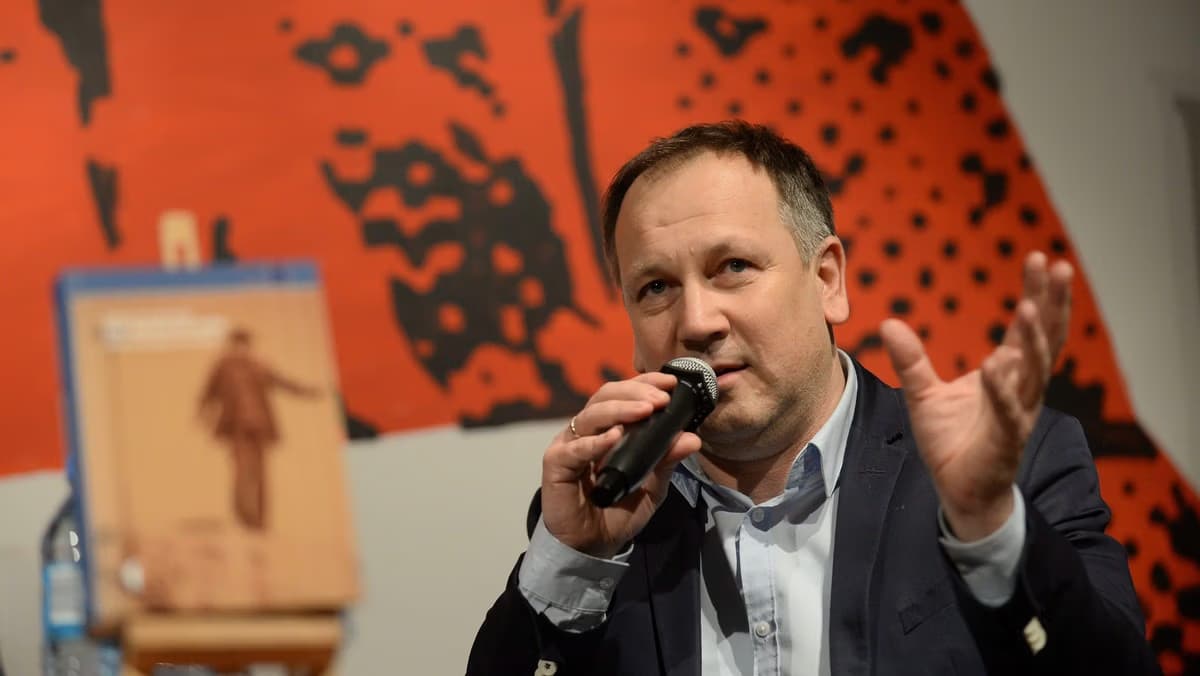 Mężczyzna w garniturze mówi do mikrofonu podczas wydarzenia wewnątrz pomieszczenia na rozmytym tle.