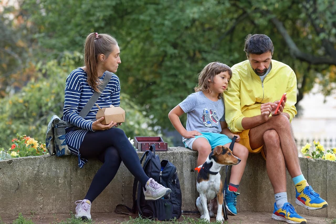 A man, woman, and child sit on a low park wall with their small dog.