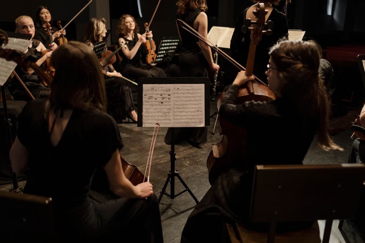 Musicians playing string instruments in a darkened orchestra, viewed from behind the cellists.
