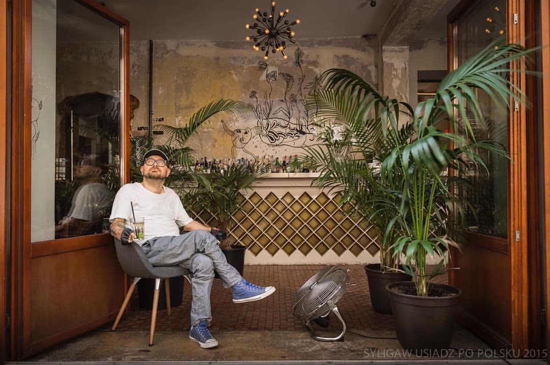 A man in a white t-shirt sits on a chair in a room with a counter and potted plants.