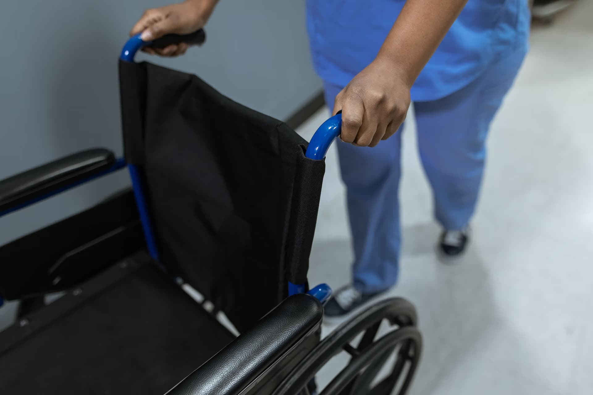 A person in blue medical scrubs pushes an empty wheelchair through a hospital hallway.